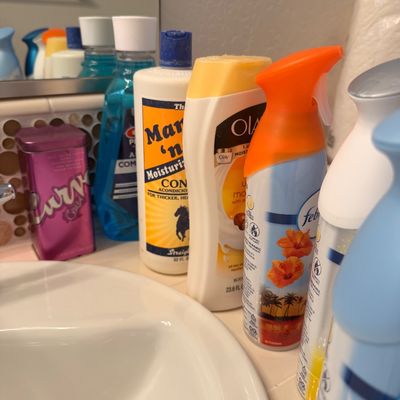 Sale Photo Thumbnail #109: Assorted personal care and household items, including Mane 'n Tail conditioner, Olay body wash, Febreze air freshener, and a Curve Crush fragrance tin, arranged on a bathroom counter.