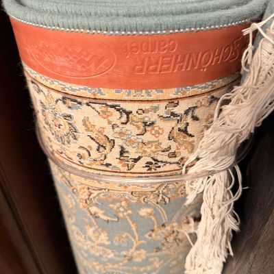 Sale Photo Thumbnail #20: Rolled Schönherr carpet featuring a light blue and cream floral pattern with fringed edges and a protective leather-look band.