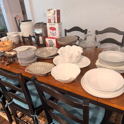 Sale Photo Thumbnail #98: Assorted kitchenware and dining items displayed on a wooden dining table, including white ceramic bowls, plates, a glass cake stand, a coffee maker, and boxed small appliances.