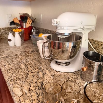 Sale Photo Thumbnail #10: White KitchenAid Artisan stand mixer with a stainless steel bowl, displayed on a granite countertop alongside vintage floral glassware and other kitchen accessories.