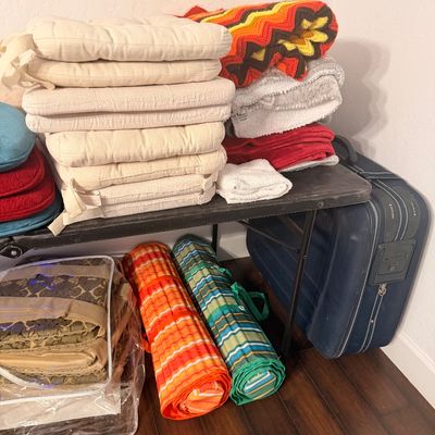 Sale Photo Thumbnail #85: Assorted household textiles including a stack of off-white chair cushions, a vintage crochet chevron blanket, towels, rolled striped mats, and a blue rolling suitcase.