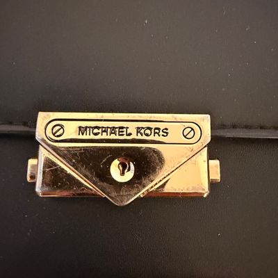 Sale Photo Thumbnail #24: Close-up of a gold-tone Michael Kors lock clasp on a black leather handbag, showing some light surface wear and scratches.