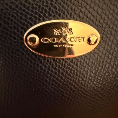 Sale Photo Thumbnail #27: Close-up of a gold-tone oval Coach New York logo plate with horse and carriage emblem on black pebbled leather.