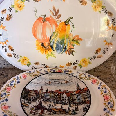 Sale Photo Thumbnail #148: Two decorative plates, featuring a large oval platter with a watercolor-style pumpkin and gourd autumn motif, and a round plate depicting a historical scene of München Marienplatz um 1700.