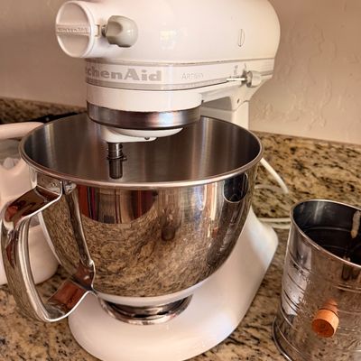 Sale Photo Thumbnail #77: White KitchenAid Artisan tilt-head stand mixer with a stainless steel bowl, shown alongside a vintage metal flour sifter.