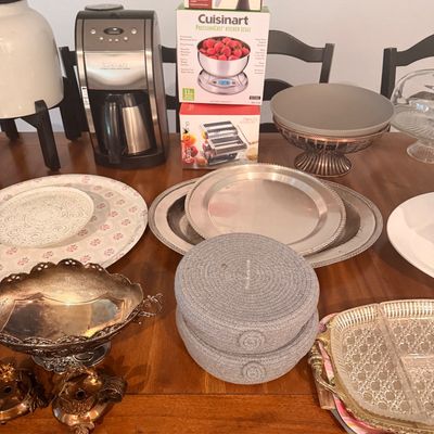 Sale Photo Thumbnail #96: Assorted kitchenware and serving pieces, including a Cuisinart coffee maker, boxed kitchen scale, silver-plated platters, and decorative bowls arranged on a wooden dining table.