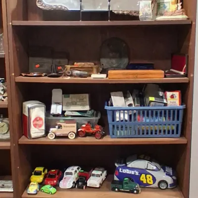 Tall wooden bookcase filled with collectibles, including vintage toy cars, a Coca-Cola napkin dispenser, a framed mirror, and various...