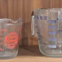 Collection of vintage glass measuring cups, including several Pyrex models with red and blue lettering, and an Anchor Hocking Oven Basics...