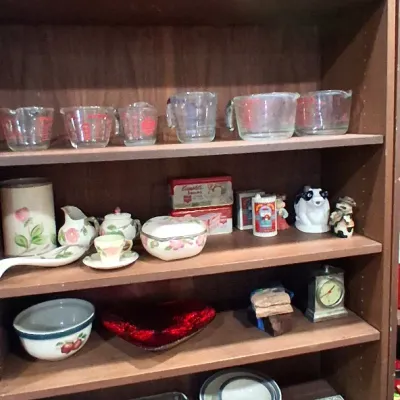 Tall wooden bookcase filled with kitchenware and collectibles, including Fire-King and Pyrex glass measuring cups, vintage ceramic...