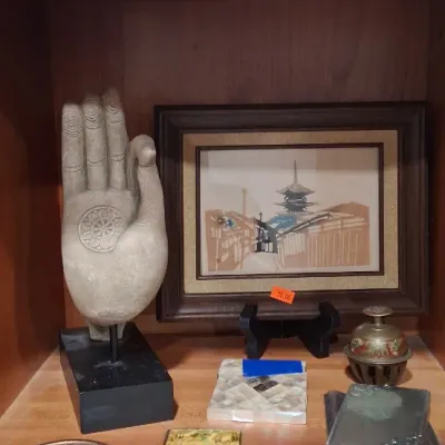 Assorted Asian-inspired decor items displayed on a wooden shelf, including a stone Buddha hand sculpture on a black stand, a framed...