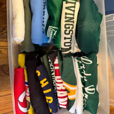 Sale Photo Thumbnail #153: Assorted athletic jerseys and t-shirts, including Livingston and Lady Devils apparel, neatly folded inside a clear plastic storage bin.