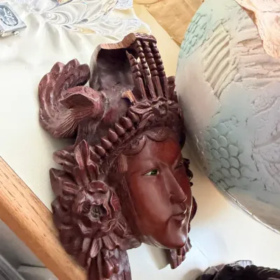 Hand-carved wooden wall mask depicting a woman's face with an ornate floral and architectural headdress, showing some damage to the...