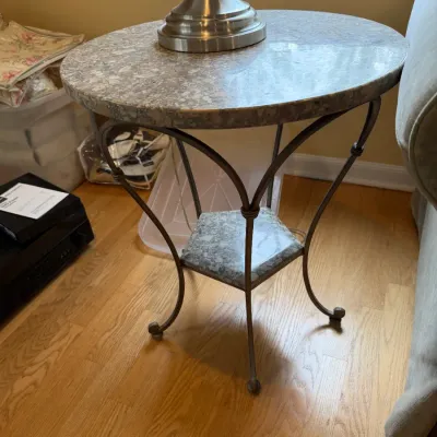 Contemporary round side table featuring a polished granite-look top and a matching lower shelf, supported by a curved dark metal frame.
