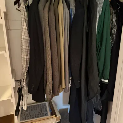 Sale Photo Thumbnail #163: Assorted men's clothing, including suit jackets, blazers, and coats, hanging in a closet alongside storage boxes.