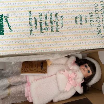 Sale Photo Thumbnail #107: Seymour Mann Connoisseur Collection porcelain doll in its original box, featuring a hand-painted porcelain head, arms, and legs, dressed in a plush white winter coat and hat with pink ribbon accents.