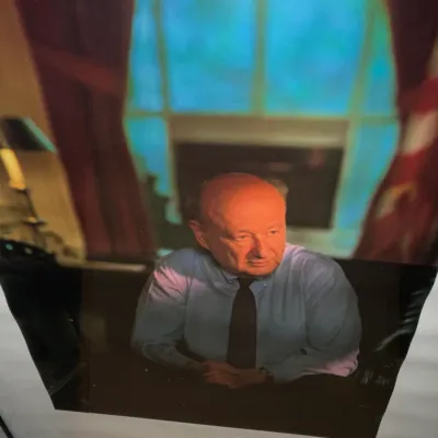 Sale Photo Thumbnail #226: Framed photographic portrait of former New York City Mayor Ed Koch, depicted sitting at a desk in front of a window with red drapes.