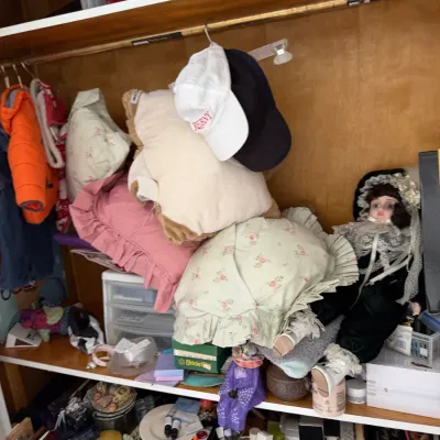Sale Photo Thumbnail #139: Assorted closet items including a porcelain doll in black and white lace attire, decorative pillows, baseball caps, and children's clothing.