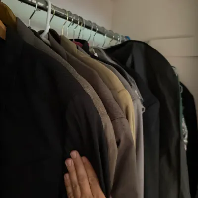 Sale Photo Thumbnail #165: Assorted men's jackets and outerwear hanging on a closet rod, including a black jacket, a tan blazer, and a dark grey coat.