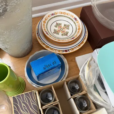 Assorted household items including a stack of patterned ceramic plates, a boxed set of glassware, a green ceramic vase, and various...
