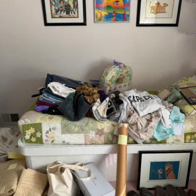 Sale Photo Thumbnail #110: Assorted household items, including framed Disney and Peter Max artwork, folded clothing, shoes, and a quilted daybed cover.