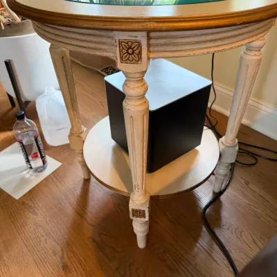 Vintage French Provincial style round side table featuring a glass insert top, distressed off-white finish with gold accents, fluted...