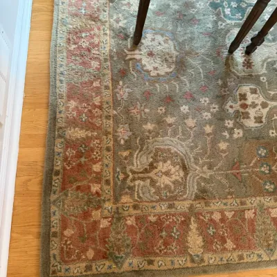 Traditional wool area rug featuring a muted green field with floral motifs and a terracotta-toned border, shown partially under a dining...
