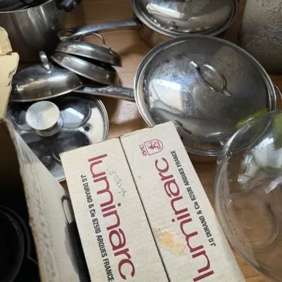 Assorted stainless steel pots and pans with lids, shown alongside a vintage Luminarc glassware box.