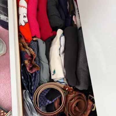 Sale Photo Thumbnail #118: Assorted clothing items, including folded shirts, sweaters, ties, and leather belts, organized inside a white dresser drawer.