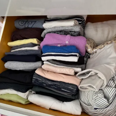 Sale Photo Thumbnail #135: Assorted women's clothing, including sweaters, t-shirts, and knit tops, neatly folded and organized inside a wooden dresser drawer.