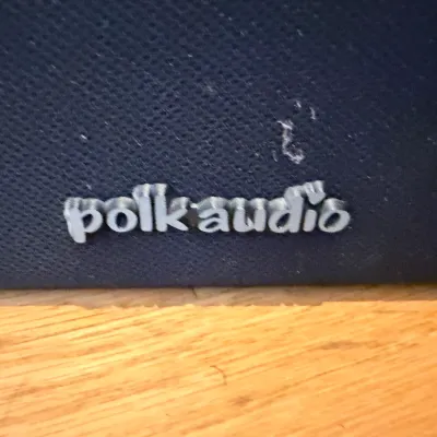 Polk Audio speaker featuring a dark blue fabric grille with a silver brand logo, showing some light wear and dust.