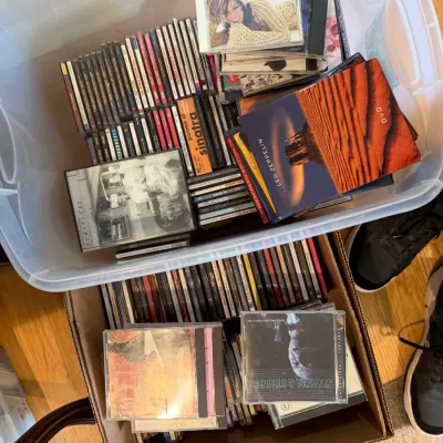 Large collection of music CDs and DVDs in plastic bins and cardboard boxes, featuring artists such as Kelly Clarkson, Frank Sinatra, and...