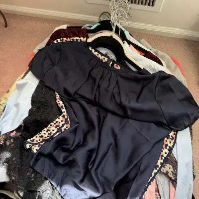 Sale Photo Thumbnail #132: A pile of assorted women's clothing on hangers, featuring a navy blue satin blouse with geometric patterned trim on top.