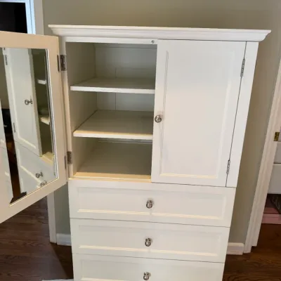Sale Photo Thumbnail #149: White wooden armoire featuring a top cabinet with shelves, a mirrored interior door, and four spacious drawers below with silver-tone knobs.