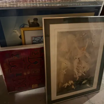 Sale Photo Thumbnail #217: Collection of framed wall art, including a large print of a woman with doves in a silver-toned frame and various other framed prints and paintings stacked behind it.
