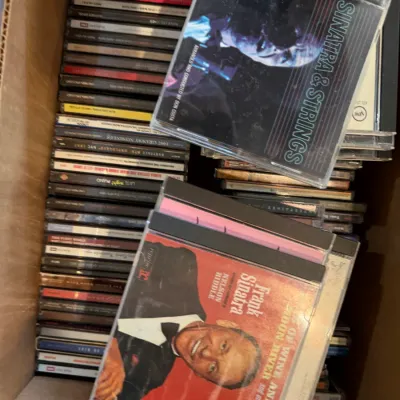 Cardboard box filled with a collection of music CDs, featuring Frank Sinatra albums including "Sinatra & Strings" and...