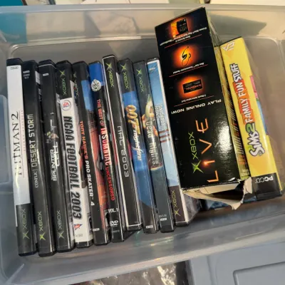 Sale Photo Thumbnail #154: Assorted vintage video games and accessories in a plastic bin, including original Xbox titles like Hitman 2, Splinter Cell, and NCAA Football 2003, alongside an Xbox Live starter kit box.