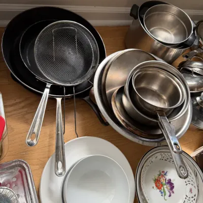 Assorted kitchenware including stainless steel pots and pans, a wire mesh strainer, white ceramic bowls, and a floral patterned plate.