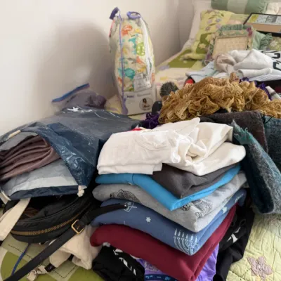 Sale Photo Thumbnail #105: Assorted clothing, textiles, and accessories, including folded sweaters, a black purse, and a decorative lace scarf, piled on a bed.