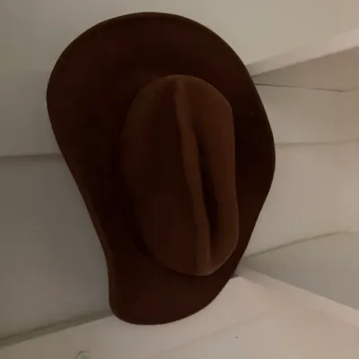 Sale Photo Thumbnail #164: Brown felt cowboy hat with a creased crown and wide brim, shown resting on a white shelf.