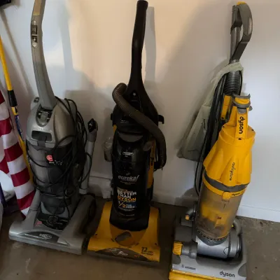 Sale Photo Thumbnail #210: Three upright vacuum cleaners including a grey Hoover WindTunnel, a black and yellow Eureka, and a yellow Dyson Root Cyclone model.