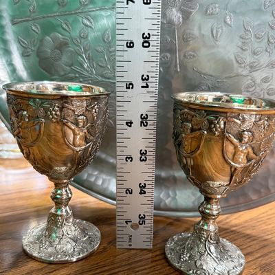 Sale Photo Thumbnail #70: A pair of vintage silver-plated goblets featuring an ornate, high-relief design of cherubs, grapevines, and musical instruments. The interiors are finished with a gold-tone wash. Each goblet stands approximately 5.5 inches tall. Shows light vintage wear a