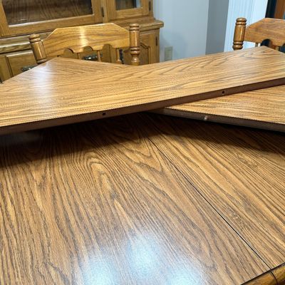 Sale Photo Thumbnail #85: A classic oak dining set featuring an oval pedestal table, two matching expansion leaves, and six spindle-back chairs. The chairs are upholstered in a dark blue textured fabric. The set shows light wear consistent with gentle use, with a clean finish on t