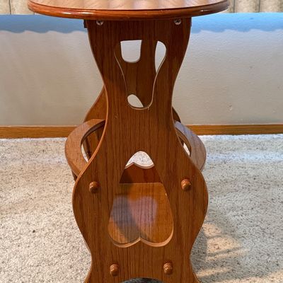 Sale Photo Thumbnail #78: A vintage wooden accent side table featuring an oval top, decorative cutout side panels, and a lower spindle-railed shelf designed to hold magazines or books. Constructed from wood with a warm honey oak finish. Shows minor surface wear, light scratches, a