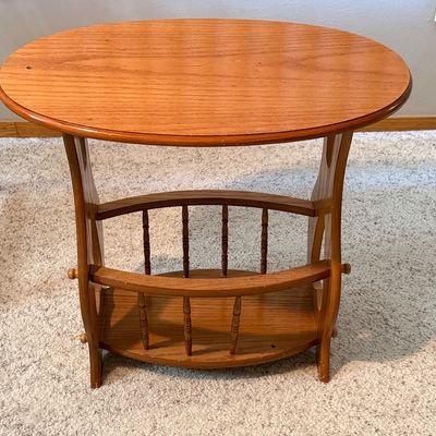 Sale Photo Thumbnail #80: A vintage wooden accent side table featuring an oval top, decorative cutout side panels, and a lower spindle-railed shelf designed to hold magazines or books. Constructed from wood with a warm honey oak finish. Shows minor surface wear, light scratches, a