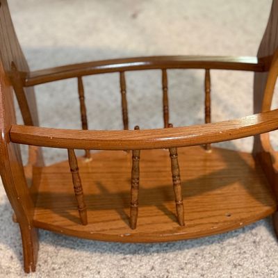 Sale Photo Thumbnail #81: A vintage wooden accent side table featuring an oval top, decorative cutout side panels, and a lower spindle-railed shelf designed to hold magazines or books. Constructed from wood with a warm honey oak finish. Shows minor surface wear, light scratches, a