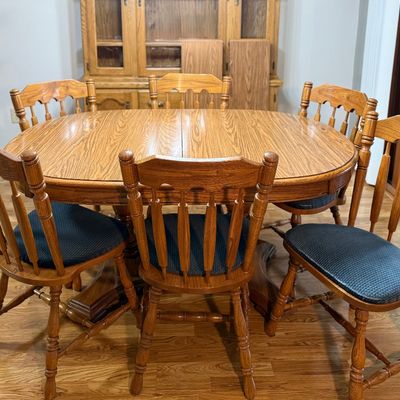 Sale Photo Thumbnail #82: A classic oak dining set featuring an oval pedestal table, two matching expansion leaves, and six spindle-back chairs. The chairs are upholstered in a dark blue textured fabric. The set shows light wear consistent with gentle use, with a clean finish on t