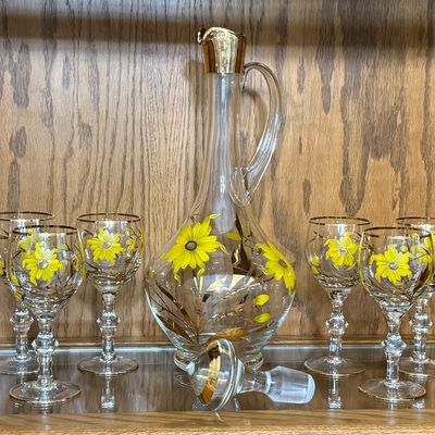Sale Photo Thumbnail #71: A beautiful mid-century vintage glassware set featuring a hand-painted yellow daisy and gold leaf design with gold-gilded rims. This 7-piece set includes one handled decanter pitcher, a matching glass stopper with a frosted ground base, and six coordinati
