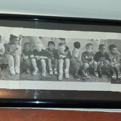 Sale Photo Thumbnail #18: Framed black and white panoramic photograph depicting a group of young children sitting in a row, housed in a simple black frame.