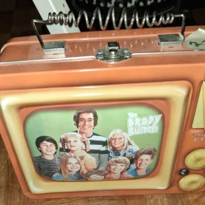 Sale Photo Thumbnail #136: Vintage-style "The Brady Bunch" collectible metal lunchbox, designed to look like a retro television set with a wire coil handle.