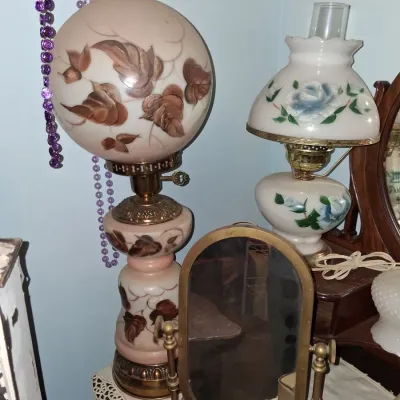 Sale Photo Thumbnail #50: Two vintage hurricane-style table lamps with hand-painted floral and leaf motifs on milk glass, shown alongside a small brass-framed vanity mirror.